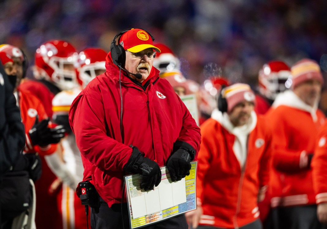 A coach in a Kansas City Chiefs jacket and hat with a headset, appears focused on a game sideline with a blurred crowd in the background.